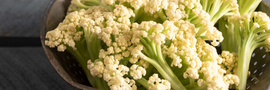The small white florets and long, pale green stems of sprouting cauliflower are displayed in a metal colander.