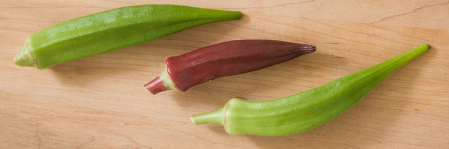 Three okra pods laid out on a wooden cutting board: two green varieties and one red variety.